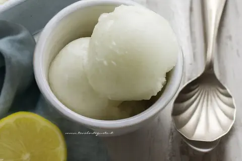 Due palline di sorbetto al limone senza albume in una ciotola bianca, con una fetta di limone e un cucchiaio decorato accanto.