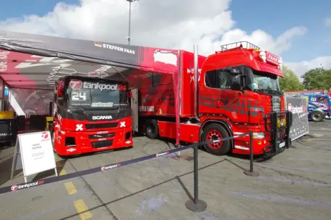 Deux camions de course rouges, prêts pour la vitesse max, sont garés sous une tente. L'un porte le numéro 24.