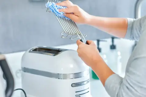 Ręce czyszczą ruszt tostera niebiesko-białą ściereczką. Dowiedz się, jak wyczyścić toster, aby był jak nowy.