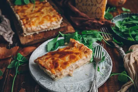 Una rebanada de tarta de cebolla dorada y cremosa, servida con rúcula fresca y un trozo de queso.