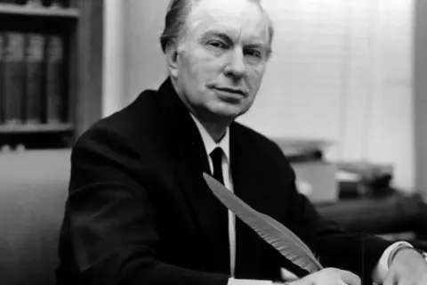 L. Ron Hubbard, twórca scjentologii, siedzi przy biurku z piórem w dłoni, gotów do pisania.