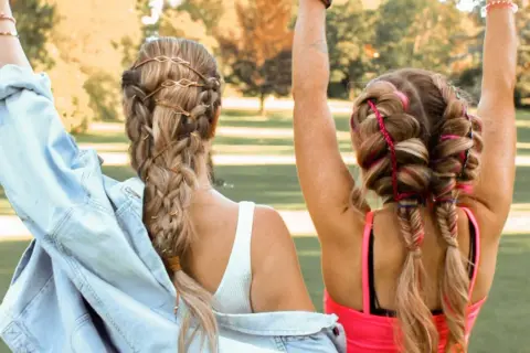 Zwei Mädchen mit geflochtenen Haaren, die sich für festival frisuren einfach eignen. Eine trägt braune Bänder, die andere bunte Fäden.