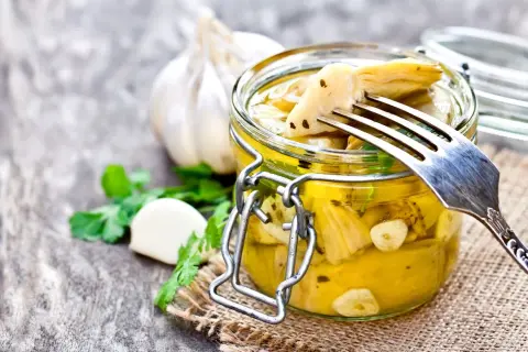 Deliziosi carciofini sott'olio fatti in casa, conservati in un barattolo di vetro con aglio e prezzemolo. Un forchettone ne solleva uno, pronto per essere gustato.