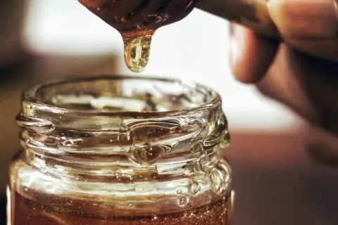 Goutte de miel doré tombant d'une cuillère dans un pot en verre. Une délicieuse alternative par quoi remplacer le sirop d'érable.