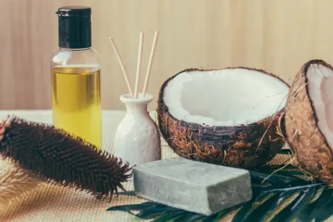 Aceite de coco para el pelo en botella junto a cocos partidos, jabón y cepillo.