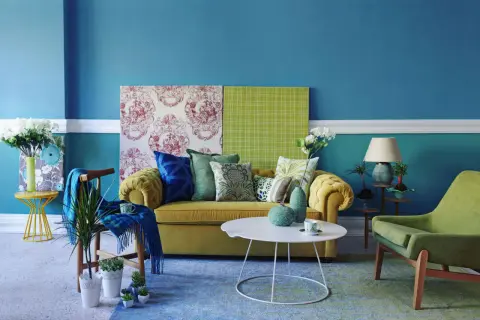 Soggiorno con divano giallo, cuscini verdi e blu, e pareti blu. Ottime idee per colorare le pareti di casa con contrasti vivaci.