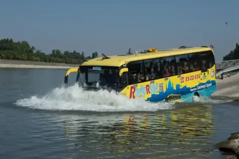 Żółty autobus "River Ride" wjeżdża do wody, tworząc fontannę. To niezwykła atrakcja turystyczna, która zabiera pasażerów na wycieczkę po Dunaju w Budapeszcie.