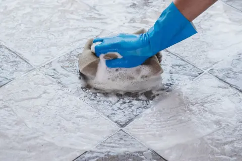 Main bleue frottant un carrelage avec une éponge, créant un voile de ciment savonneux.