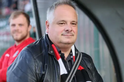 Prezes Polonii Warszawa w skórzanej kurtce i szaliku klubu, z uśmiechem na twarzy.