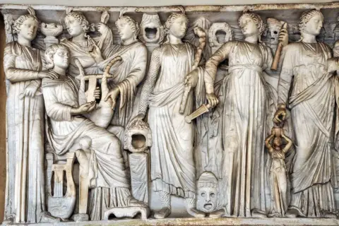 Kamienna płaskorzeźba przedstawia grupę kobiet, w tym Demeter i Korę, otoczone instrumentami muzycznymi i symbolicznymi przedmiotami.