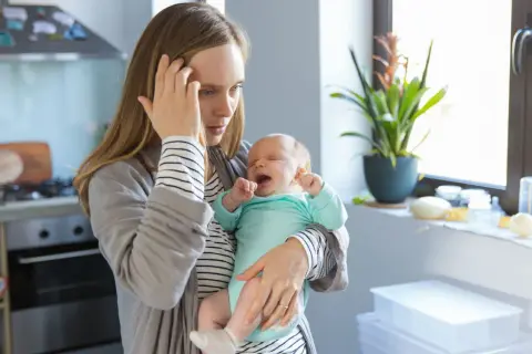 Erschöpfte Mutter hält weinendes Baby. Der 12 wochen schub macht ihr zu schaffen. Sie hält sich die Stirn.