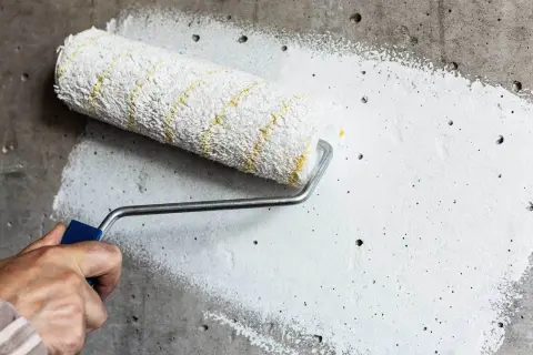 Dłoń trzymająca wałek malarski z białą farbą na szarej, chropowatej ścianie.