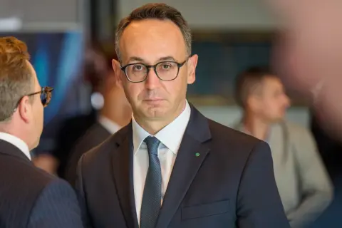 Minister infrastruktury w okularach i garniturze, z zieloną koniczynką na klapie marynarki, rozmawia z kimś na tle innych osób.
