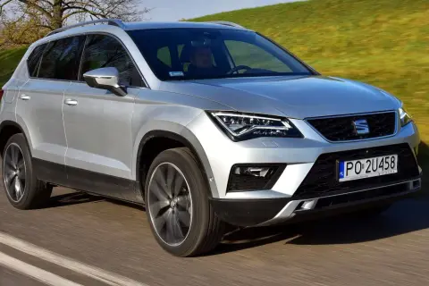Srebrny Seat Ateca w ruchu. Kierowca za kierownicą, zielone wzgórza w tle. Wiele osób szuka seat ateca opinie przed zakupem.