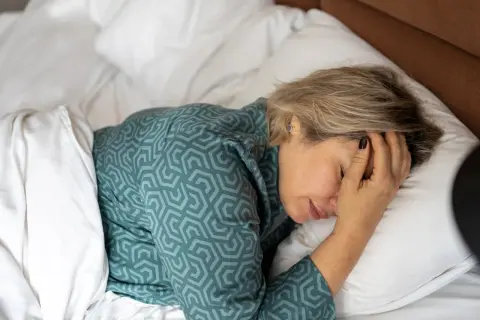 Een vrouw ligt in bed, met haar hand op haar voorhoofd. Ze vraagt zich af: waarom voel ik me zo moe?