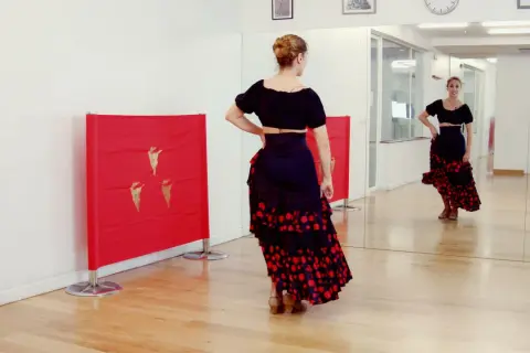 Bailarina con falda flamenca y top negro, practicando una rumbita flamenca frente a un espejo y paneles rojos con detalles dorados.