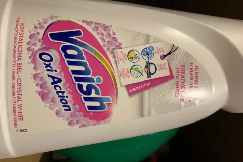 Butelka płynu Vanish Oxi Action Crystal White z etykietą i zawieszką informującą o działaniu.