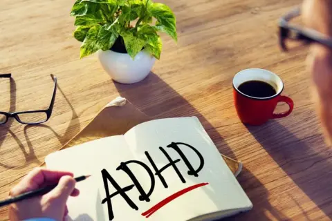 Osoba zmagająca się ze spektrum ADHD pisze w zeszycie. Obok leżą okulary, filiżanka kawy i roślina.