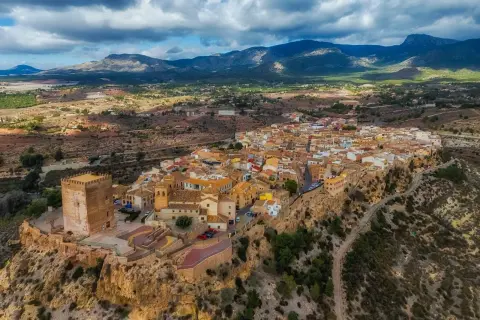 Pueblo blanco en un acantilado con castillo, ideal para saber qué ver en Murcia y alrededores. Paisaje montañoso al fondo.