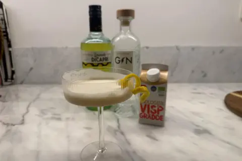 En krämig limoncello drink med citronskal, bredvid flaskor med limoncello, gin och vispgrädde.