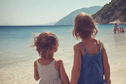 Dwie dziewczynki trzymają się za ręce na plaży, patrząc na morze. W tle widać góry i innych ludzi kąpiących się w wodzie. To idealne wakacje w Albanii z dziećmi.