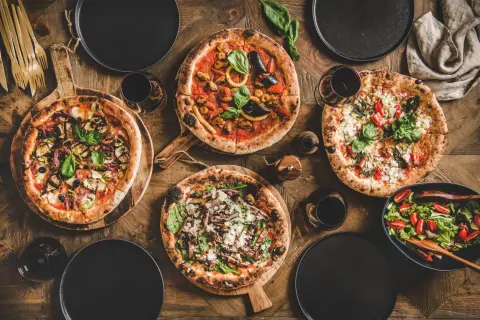 Vier Pizzen, ein Salat und Rotwein auf Holztisch. Die Frage welcher Wein zu Pizza wird hier beantwortet.