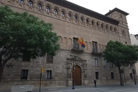 Fachada del palacio de los Luna en Zaragoza, con sus arcos y balcones, flanqueado por árboles verdes.
