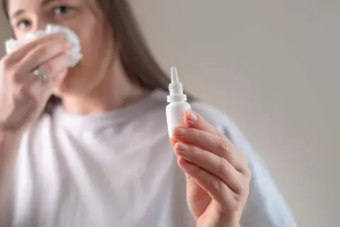 Kobieta z chusteczką przy nosie trzyma w dłoni małą buteleczkę z kroplomierzem.