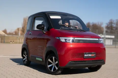 Czerwony, mały samochód elektryczny z kobietą za kierownicą, na tle placu i nieba.