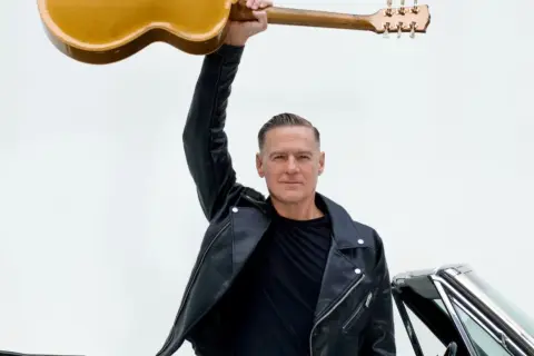 Bryan Adams w skórzanej kurtce unosi gitarę nad głową, gotowy na koncert. Obok klasyczny samochód.