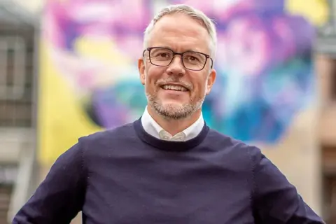 Niclas Hermansson ler glatt framför en färgglad väggmålning.