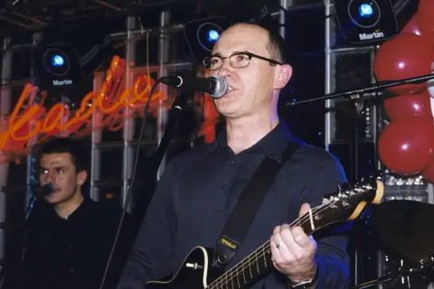 Wokalista z gitarą śpiewa na scenie, a w tle neonowy napis. Czas na koncert, a kiedy przyjdzie na ciebie czas tekst.