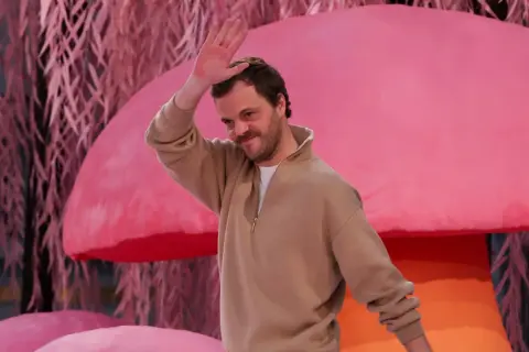 Matthieu Blazy saluta con la mano, sorridendo, su uno sfondo rosa con elementi che ricordano funghi e piante.