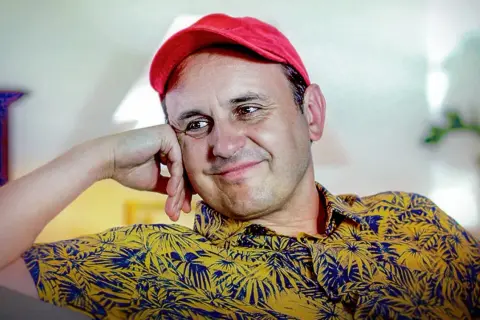 Uśmiechnięty Adam Woronowicz w czerwonej czapce i żółtej koszuli w liście. Wiele jego filmów zapisało się w pamięci widzów.