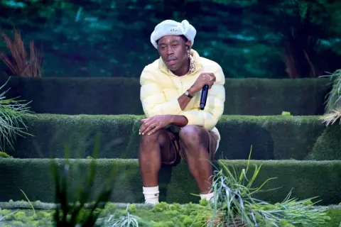 Igor, czyli Tyler, The Creator, siedzi na scenie w żółtej kurtce i jasnoniebieskiej czapce, trzymając mikrofon.