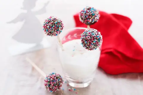 Färgglada cake pops med strössel står i ett glas med mjölk. En röd duk och en ängel i bakgrunden skapar en festlig stämning.