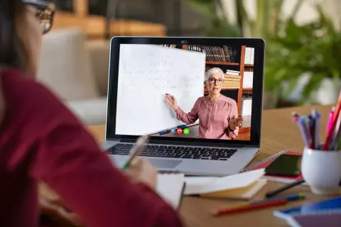 Nauczycielka prowadzi lekcję online na laptopie, wskazując na tablicę z równaniami.