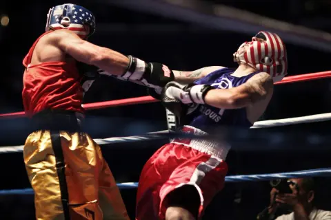 Boxer in red shorts lands a punch to the solar plexus of his opponent in red and white striped headgear.