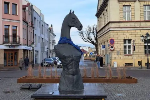 Pomnik z głową konia i niebieską szarfą, przypominający herb. W tle kamienice i ulica.