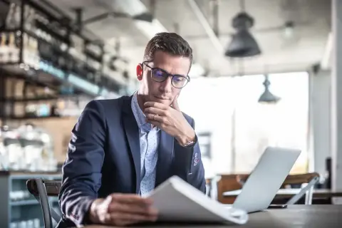 Mężczyzna w okularach i garniturze analizuje dokumenty przy laptopie w kawiarni.