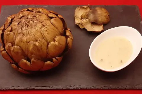 Un artichaut entier, cuit à la cocotte minute, servi avec sa sauce et quelques feuilles détachées sur une ardoise.