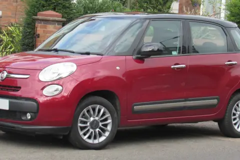 Czerwony Fiat 500 L z czarnym dachem zaparkowany przy ceglanym murze i zieleni.