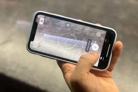 Dłoń trzyma smartfon, na ekranie którego działa aplikacja miarka. Wirtualna linijka mierzy odległość na powierzchni.