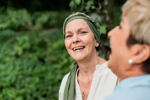 Zwei Frauen lachen im Freien. Die vordere Frau trägt ein Kopftuch und wirkt durch psychoonkologische Beratung gestärkt.