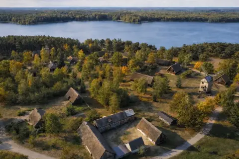 Wdzydze Kiszewskie: skansen z drewnianymi chatami i wiatrakiem nad jeziorem, otoczony jesiennym lasem.