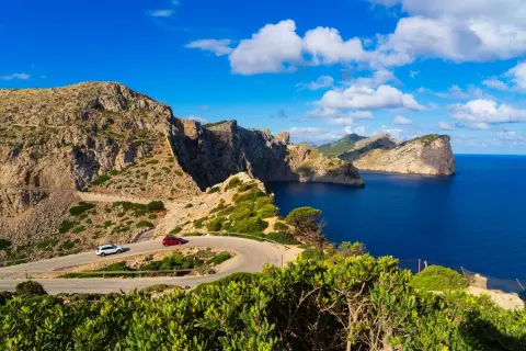 Coche rojo y blanco serpentean por la **Cap de Formentor route**, con acantilados escarpados y el mar azul intenso.
