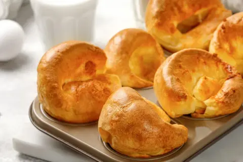 Goldbraune Yorkshire Pudding im Muffinblech, bereit für ein köstliches Rezept. Daneben ein Ei und ein Glas Milch.
