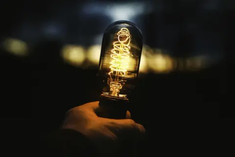 Dłoń trzyma żarówkę 1500 lumenów, której żarnik świeci ciepłym, żółtym światłem. Tło jest ciemne, z rozmytymi, jasnymi plamami.