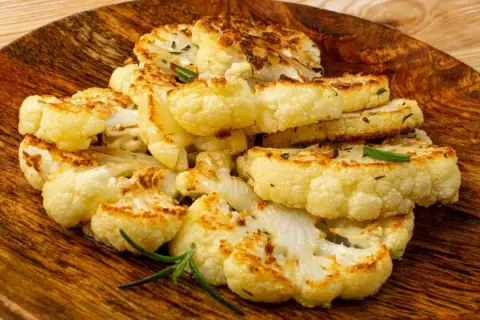 Tranches de chou-fleur entier au four, dorées et savoureuses, garnies de romarin frais sur une assiette en bois.