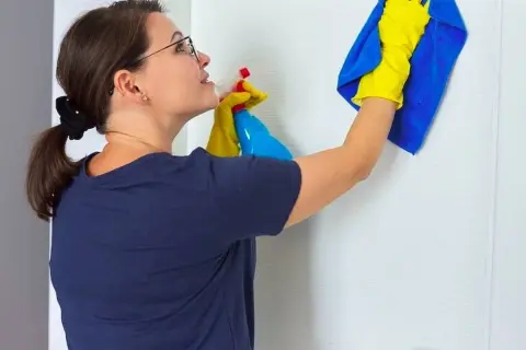 Mujer con guantes amarillos y paño azul limpiando puerta lacada en blanco con spray.